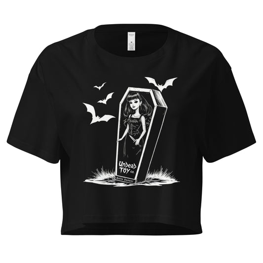 Black crop top with a graphic design of a doll named Dolly Dagger holding a dagger  in a coffin doll box with bats flying around. 'Undead Toy co' is written on the toy box.